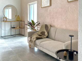 A modern living room with a light grey sofa and a small black side table. A decorative plant and mirror are in the background, next to a console table with vases.