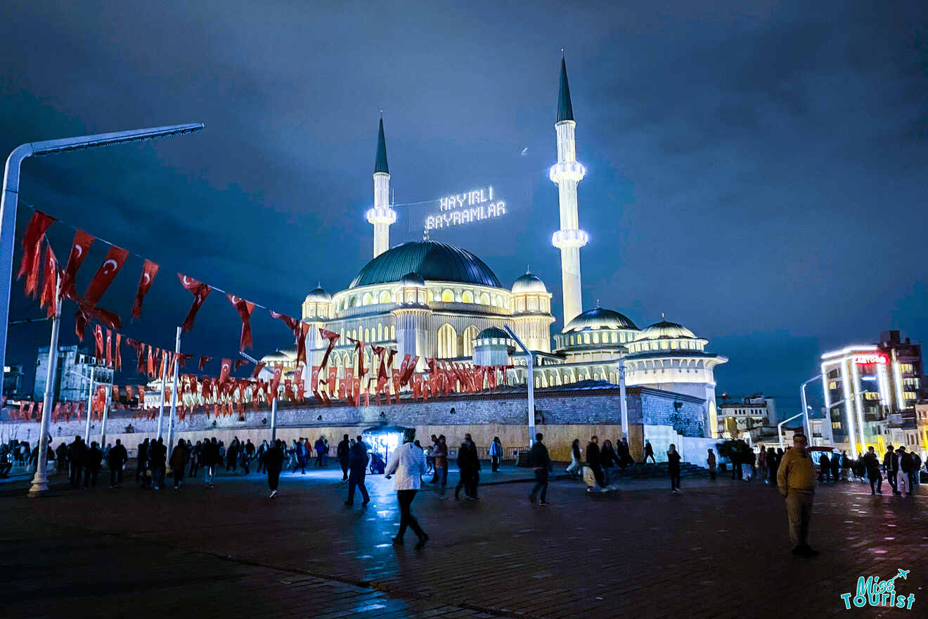 Night view of a lit mosque with two minarets, surrounded by people and red flags, under a cloudy sky.