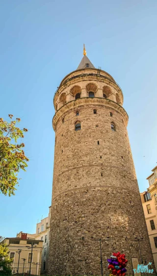 Galata Tower in Istanbul, with a conical roof and ancient stone structure, stands against a clear blue sky. A small cluster of colorful balloons is visible at the bottom right.