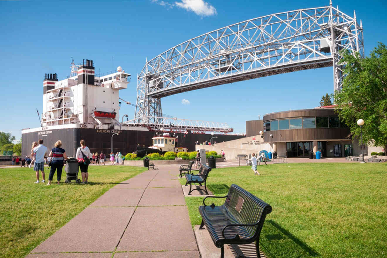 A large ship passes under a raised lift bridge as people stand nearby on a grassy area with benches.