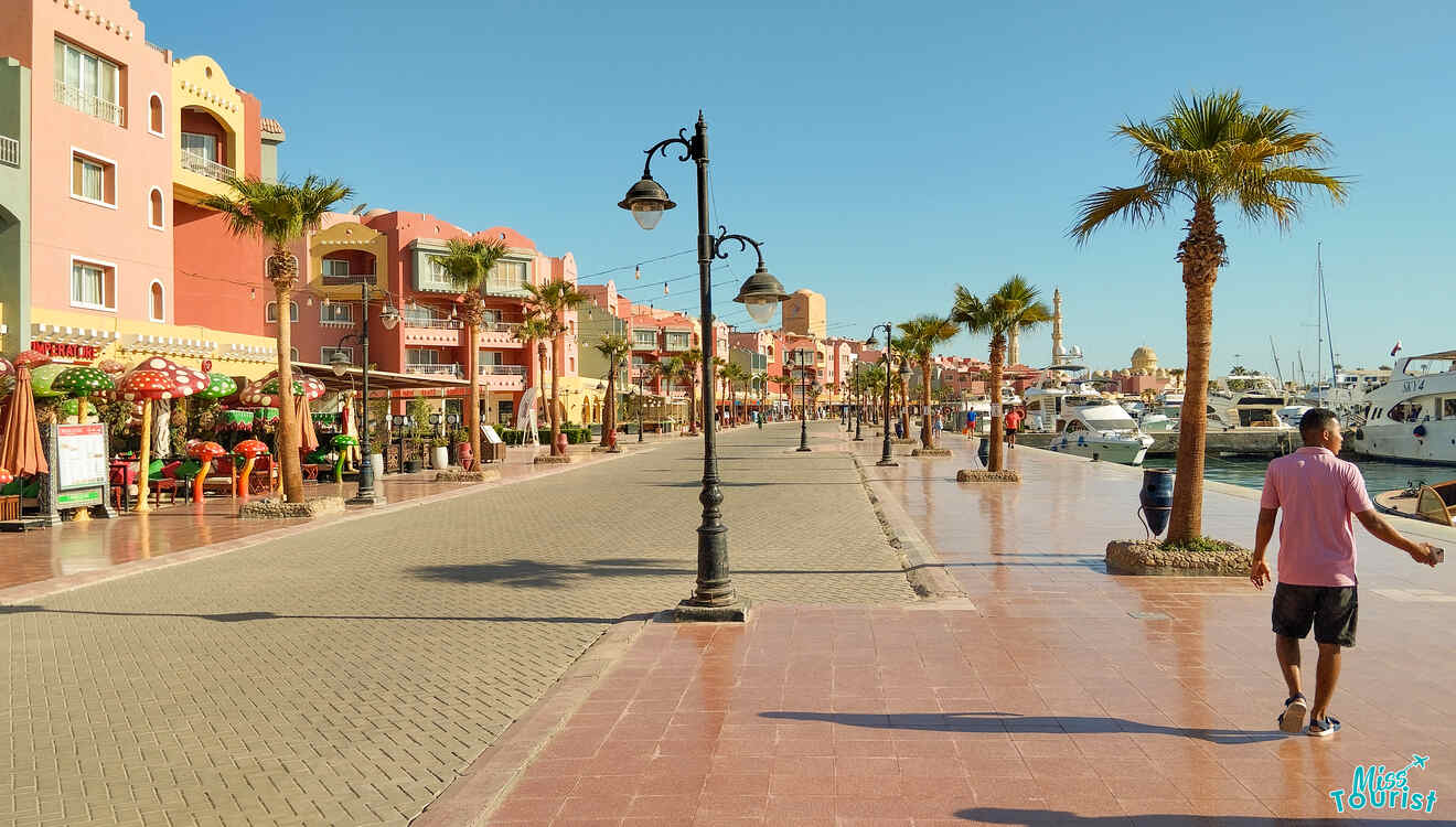 A sunny promenade with palm trees, colorful buildings on the left, and boats docked on the right. A person walks along the path.