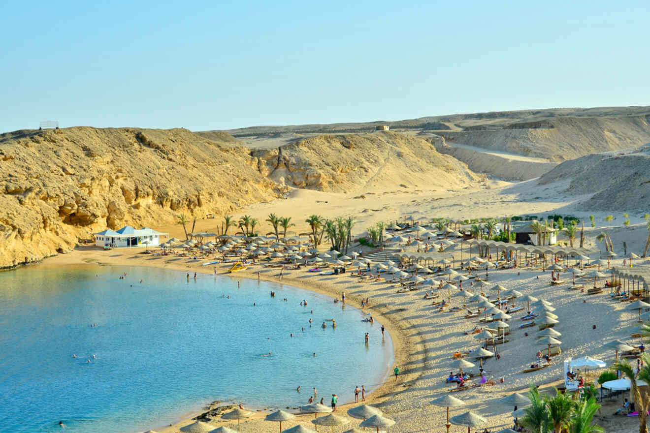 Beach with umbrellas and loungers along a sandy shoreline, surrounded by rocky hills. People are swimming and sunbathing.