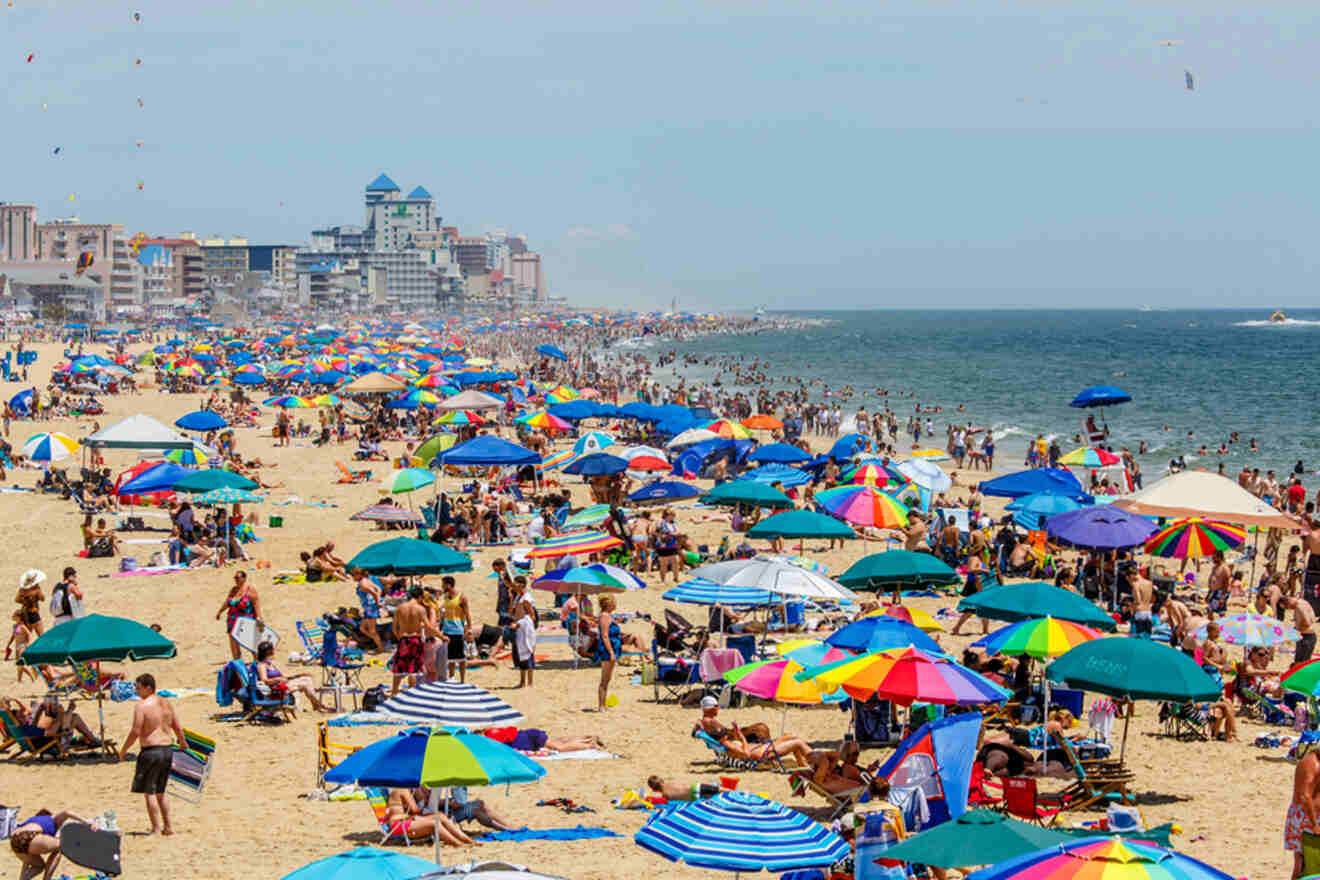 Crowded beach with colorful umbrellas, people sunbathing, and buildings in the background.