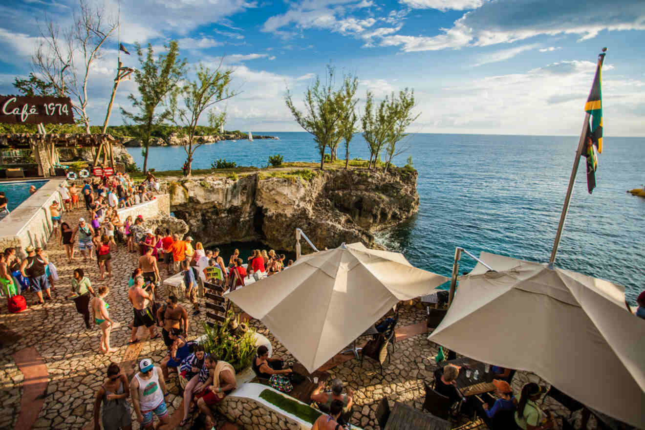 Crowded seaside café with people gathered near rocky cliffs and the ocean. Umbrellas shade tables, and a flag waves in the breeze.