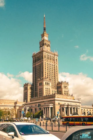 Tall building with a spire, resembling classic Soviet architecture, under a blue sky. Public transportation and cars are in the foreground.