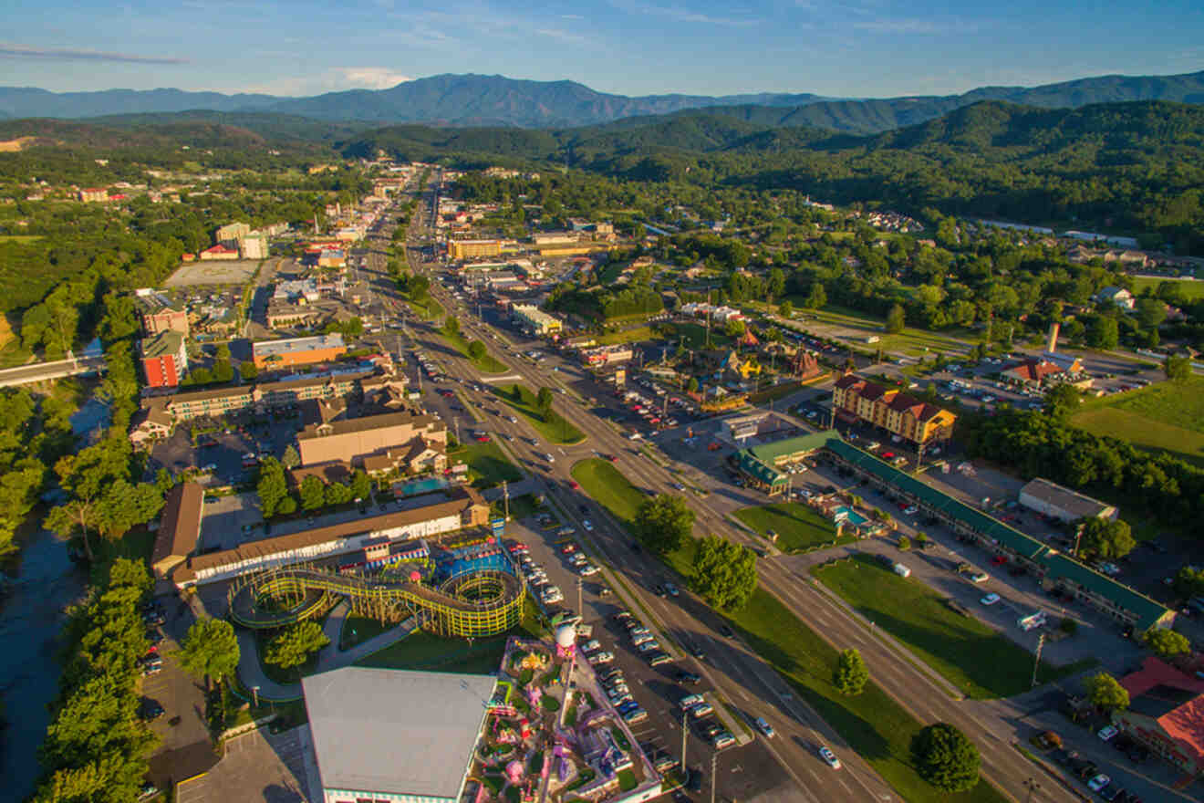 Aerial view of a town with busy roads, colorful amusement park rides, and surrounding green hills under a blue sky.