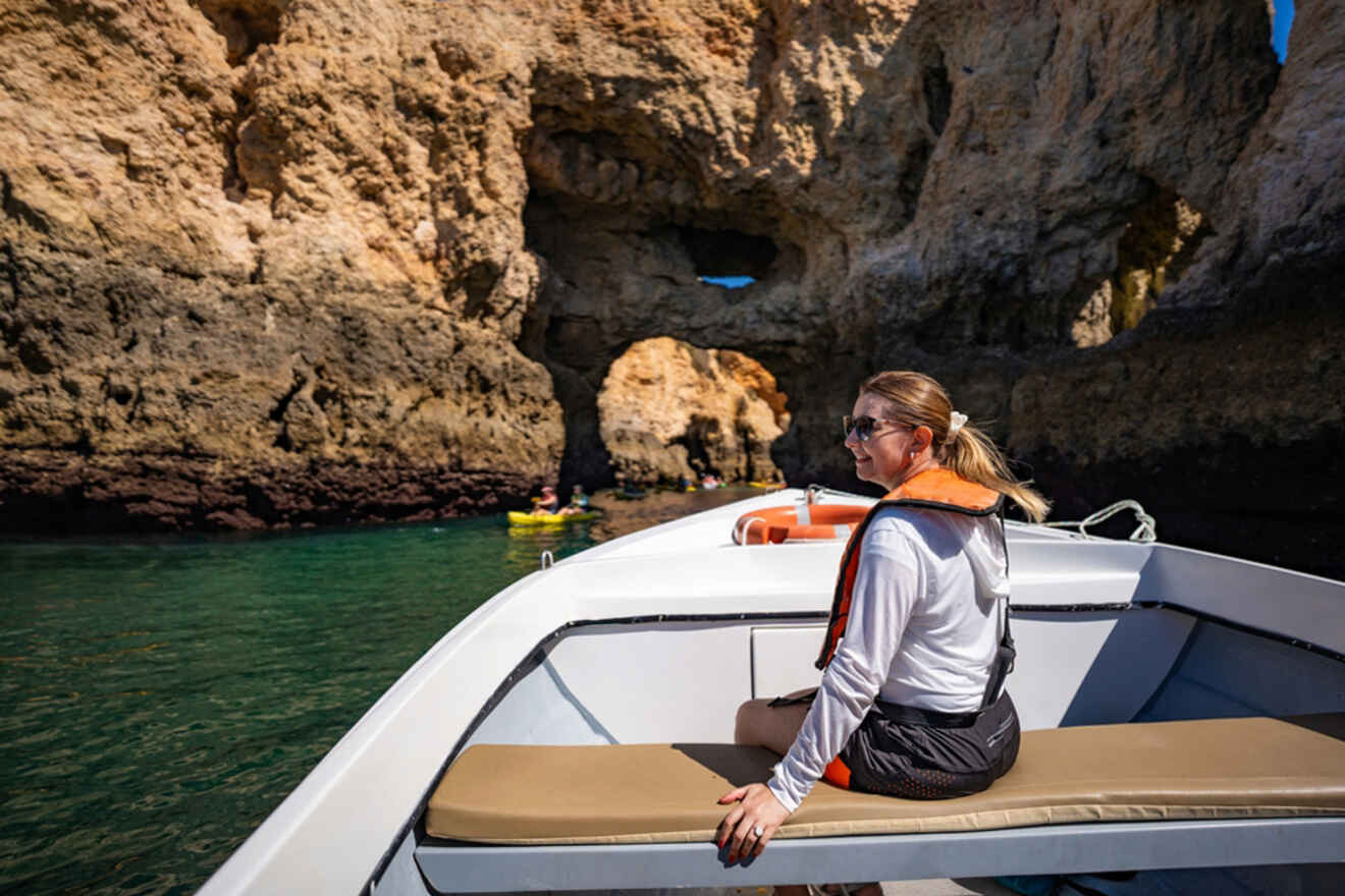 A person sits on a boat in front of the rocky cliffs and clear waters of Lagos, Portugal, with a kayak nearby.