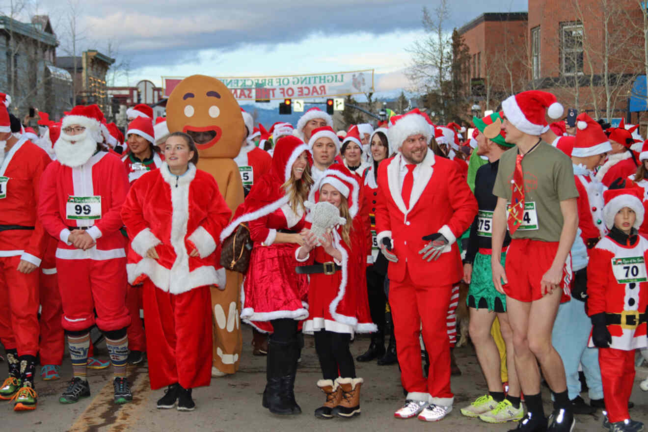 A large group of people dressed in Santa and holiday-themed outfits participate in a festive outdoor event, with a banner visible in the background.