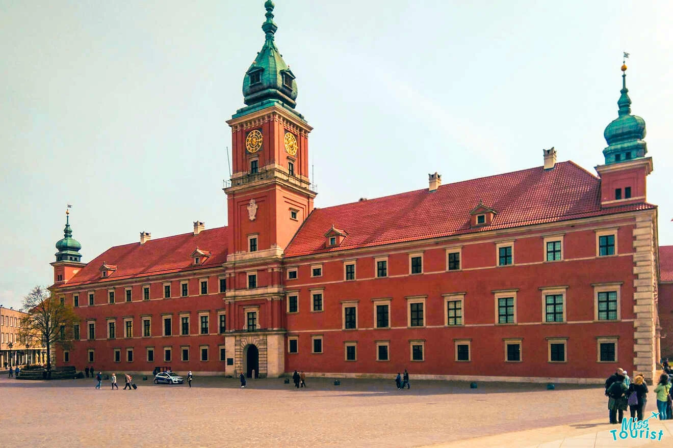 A large red-brick building with green-roofed towers stands on a cobblestone square. A few people walk in the foreground under a clear sky.