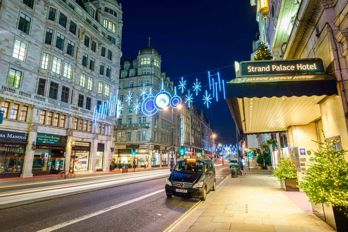 A city street at night with festive lights hanging above. Strand Palace Hotel is on the right, and buildings line the road. A black car is parked by the curb.