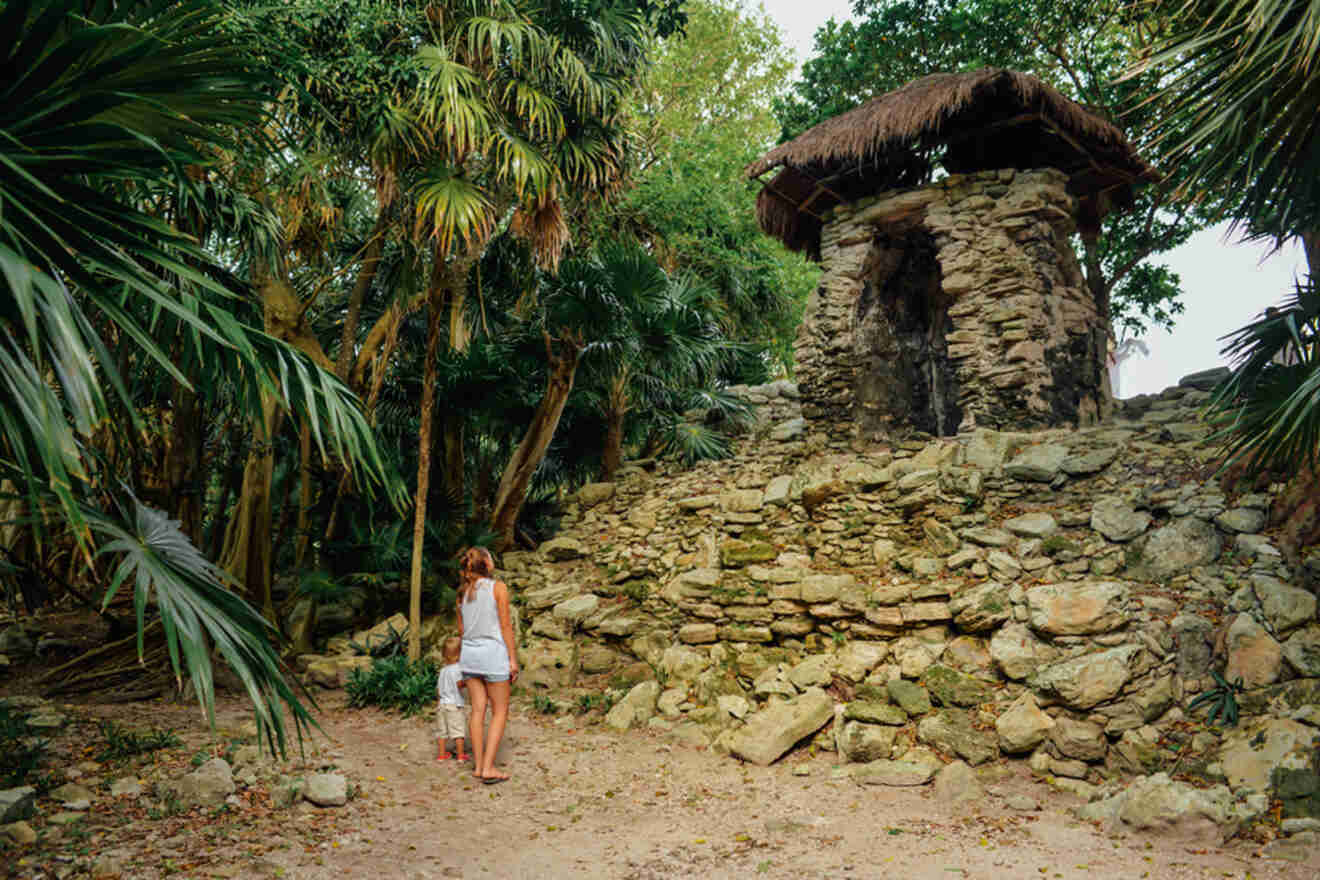 A person stands near an ancient stone structure surrounded by lush greenery and palm trees.