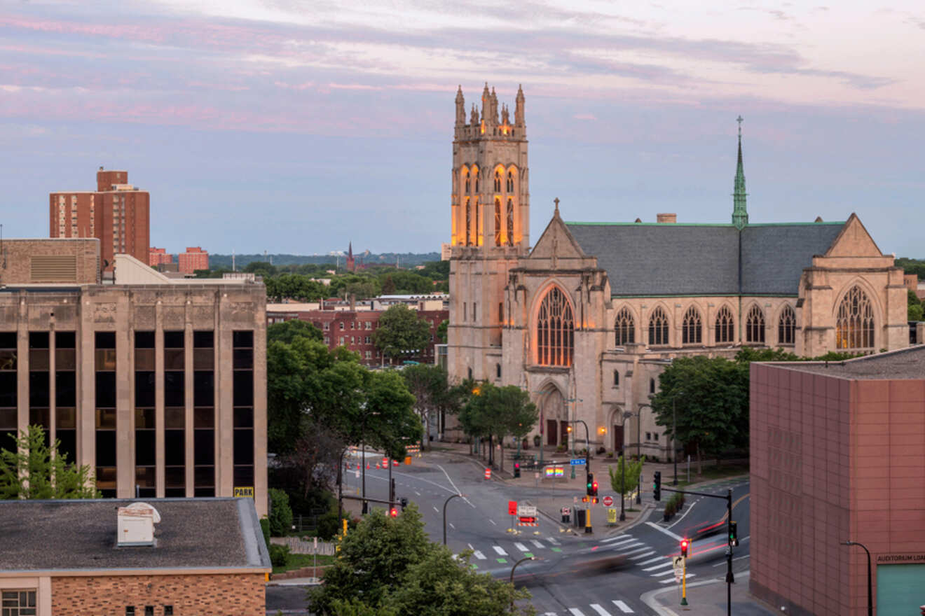 Aerial view of a cityscape featuring a large cathedral at the center, surrounded by buildings and a street with sparse traffic. Sky is partly cloudy with a soft pastel hue.