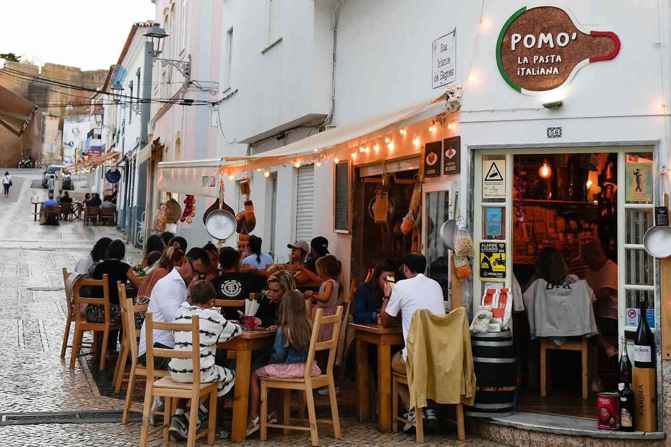 People dining outside a restaurant on a narrow street with tables along the sidewalk. The restaurant sign reads "Pomo, La Pasta Italiana.