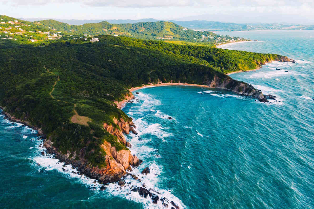 Aerial view of a coastal landscape with lush green hills, rocky cliffs, and waves crashing onto the shoreline, set against a blue ocean and a partly cloudy sky.