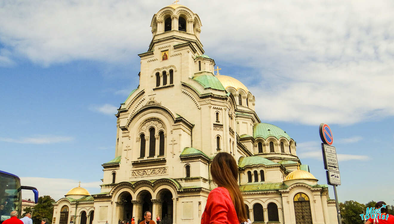 A large cathedral with domes under a cloudy sky. The writer of the post in a red jacket walks in front.