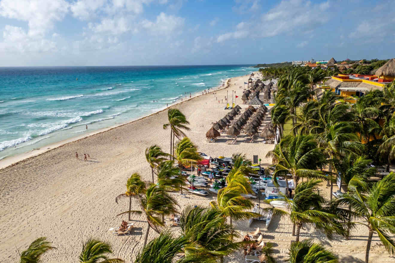 Aerial view of a sandy beach with palm trees, lounge chairs, and umbrellas near turquoise ocean waves. People stroll along the shoreline on a sunny day.