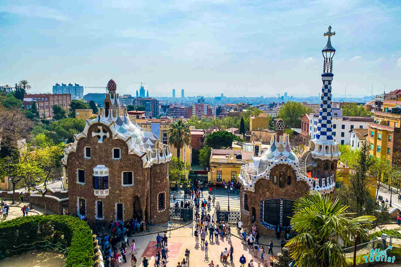 Aerial view of Park Güell entrance in Barcelona, featuring two unique buildings with tile roofs, surrounded by greenery and crowds of visitors below. Cityscape in the background.