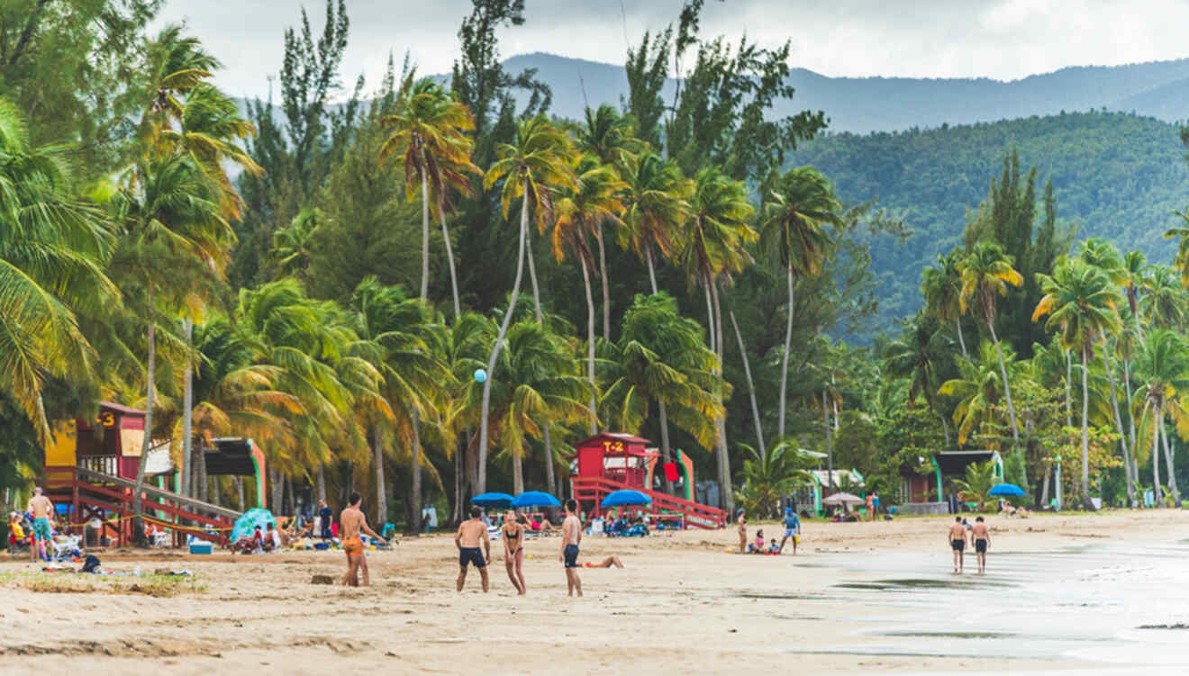 People relax and stroll on a tropical beach with palm trees, blue umbrellas, and lifeguard stations, set against a backdrop of green hills.