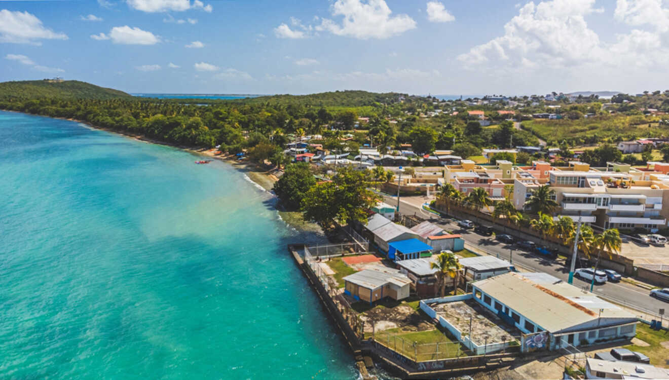 Aerial view of a coastal town with turquoise water, buildings, and lush greenery under a partly cloudy sky.