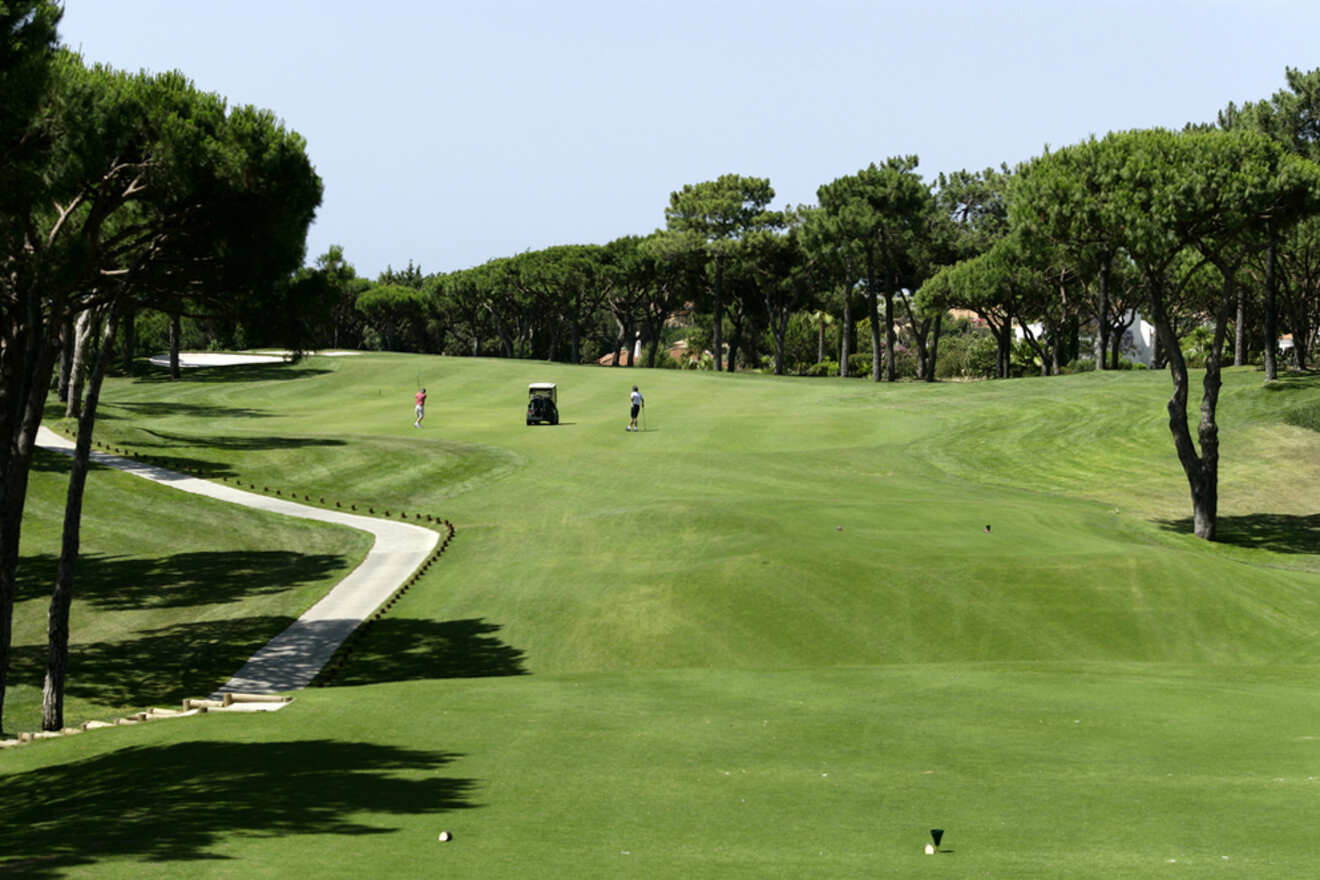 Amidst the lush green fairways of a golf course in Lagos, two people stroll leisurely near a golf cart, surrounded by tall trees under Portugal's clear sky.