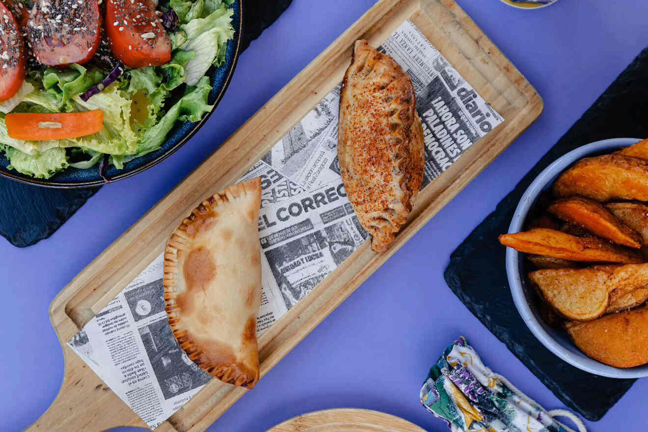 A wooden board with two empanadas on newspaper, surrounded by a salad, a bowl of potato wedges, and a small wrapped item on a purple surface.