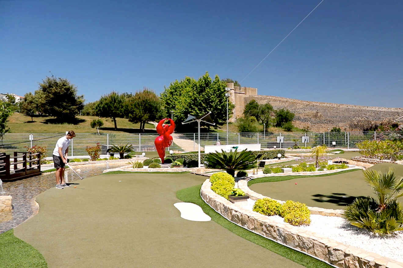 A person enjoys mini golf on a sunny day in Lagos, Portugal, surrounded by lush greenery and a large red statue in the background.