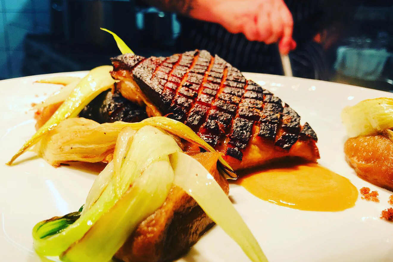 Grilled meat with a crisscross char pattern served with roasted vegetables and a sauce on a white plate. A person's hand is visible in the background.