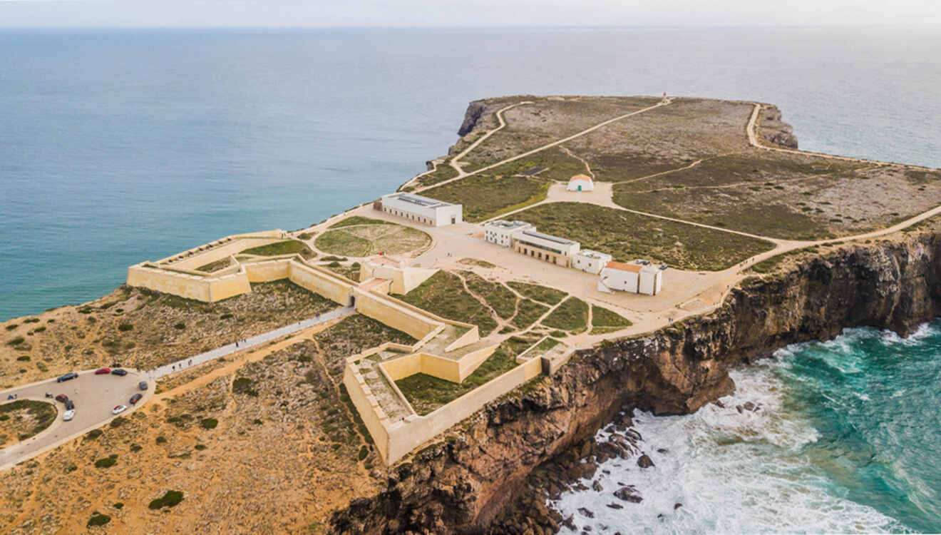 Aerial view of a historic fort on a cliff in Lagos, Portugal, overlooking the ocean. The fort is surrounded by rugged terrain with pathways and structures. Waves crash against the rocky shoreline below.