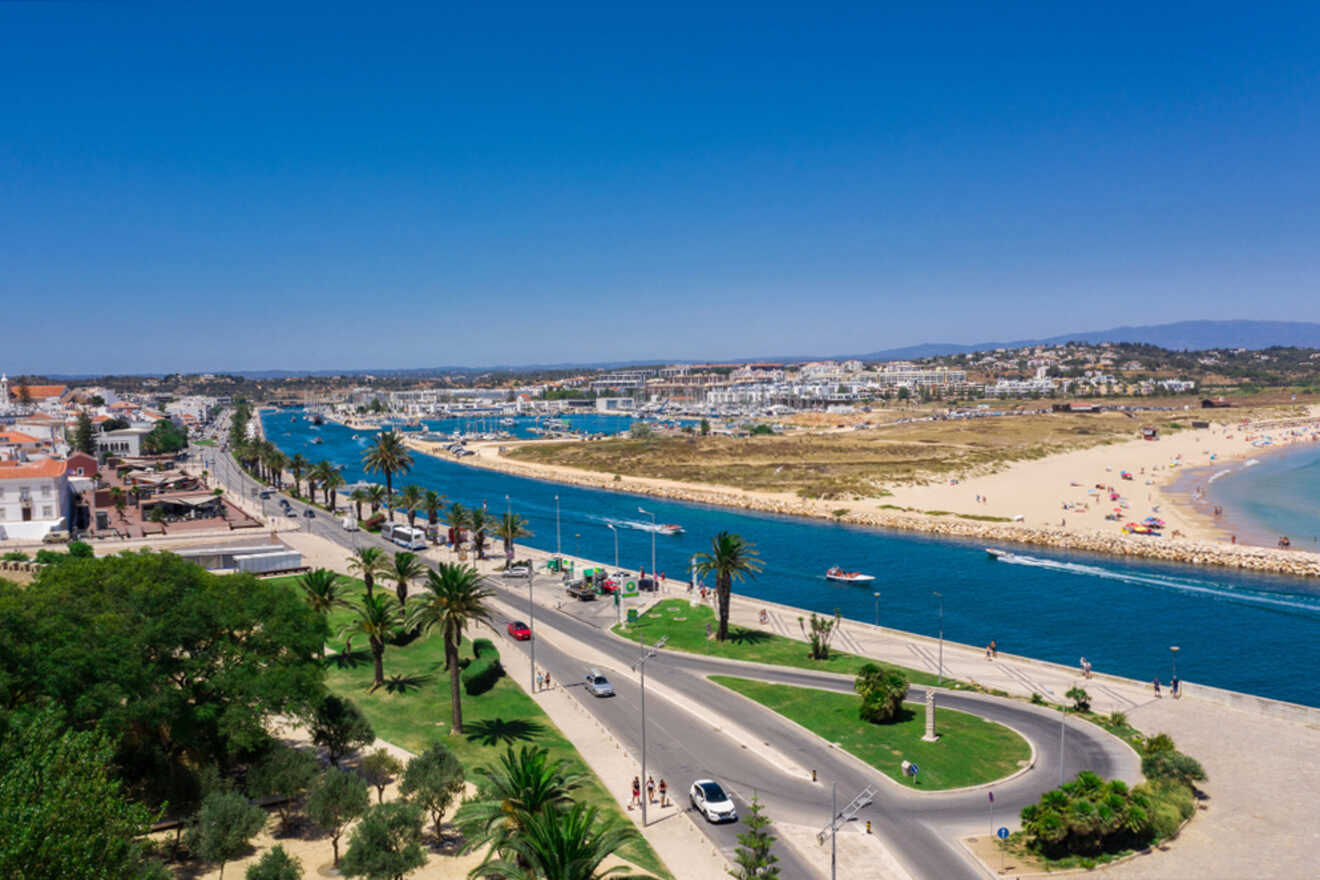 Aerial view of the coastal town of Lagos, Portugal, featuring a river, sandy beach, and marina. Palm-lined streets and charming buildings are visible under a clear blue sky.