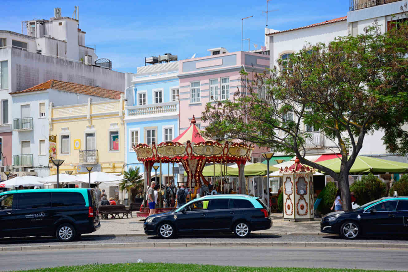 A street scene in Lagos, Portugal, with pastel-colored buildings and a charming carousel. Outdoor seating invites passersby to unwind, while black vehicles are parked in the foreground, adding a contemporary contrast to the picturesque setting.