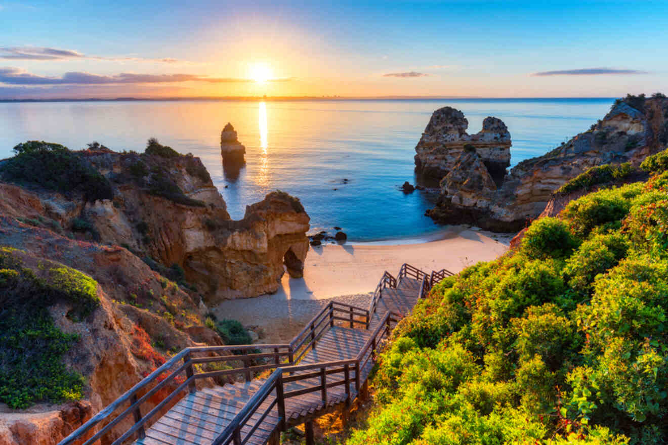 Wooden stairs lead to a beach surrounded by rocky cliffs at sunrise in Lagos, Portugal, with vibrant greenery and a calm sea.