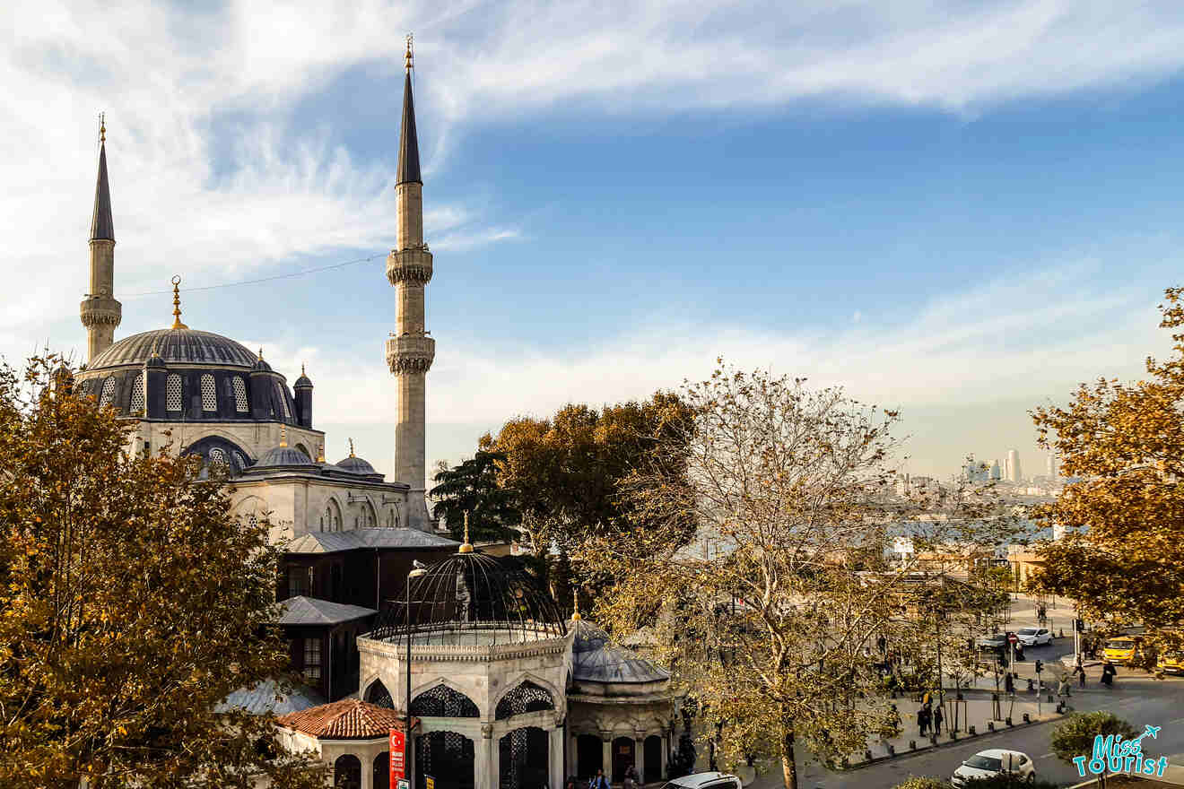 A mosque with two minarets stands against a blue sky. Trees with autumn leaves surround the building. People walk nearby, and a cityscape is visible in the distance.