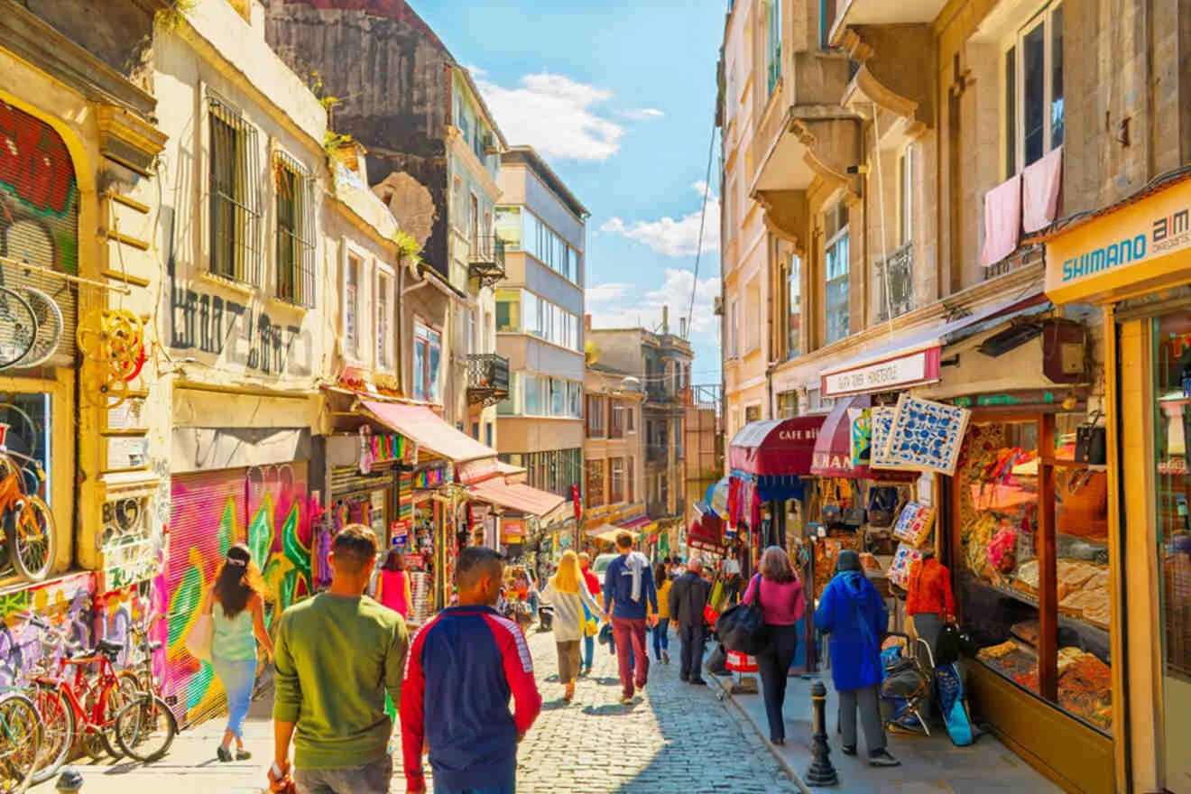 A bustling street scene with people walking along cobblestones, flanked by colorful buildings, shops, and street art under a bright blue sky.