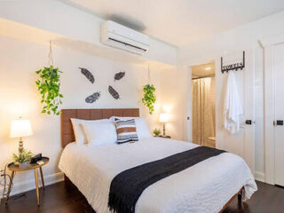 A cozy bedroom with a neatly made bed, two side tables with lamps, hanging plants, decorative wall feathers, and an open door revealing a bathroom with a towel rack.