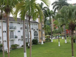 A white multi-story building with balconies is surrounded by palm trees on a well-maintained lawn.