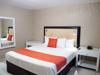 A hotel room with a large bed, featuring white and orange bedding. Two nightstands with a lamp and phone are on either side. A mirror is mounted on the beige wall to the left.