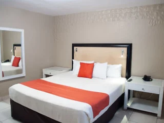A hotel room with a large bed, featuring white and orange bedding. Two nightstands with a lamp and phone are on either side. A mirror is mounted on the beige wall to the left.