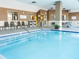 Indoor swimming pool with clear blue water, surrounded by a tiled deck area. Chairs and tables are positioned along the wall. A staircase with handrails leads into the pool.