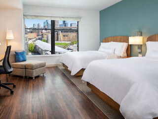 A hotel room with two beds, wooden flooring, a desk with a chair, a cityscape view from the window, and teal accent wall.