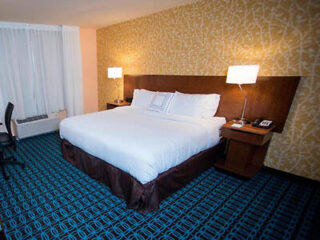 Hotel room with a king-sized bed, wooden headboard, two bedside tables with lamps, patterned blue carpet, and a decorative yellow wall. A desk and chair are on the left.