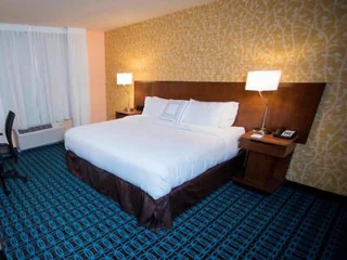 Hotel room with a king-sized bed, wooden headboard, two bedside tables with lamps, patterned blue carpet, and a decorative yellow wall. A desk and chair are on the left.