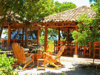 A rustic outdoor seating area with wooden chairs and tables under shingled pavilions surrounded by lush greenery.