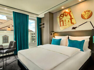 Modern hotel room with a double bed, turquoise pillows, a large window with teal curtains, wall art, and a small seating area with a table and chair.