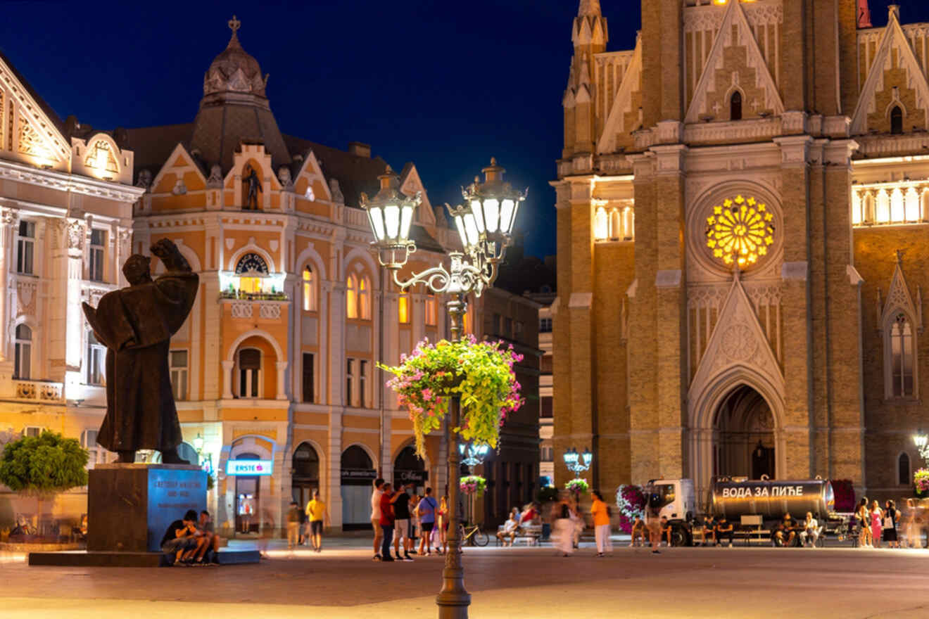 A city square at night featuring a lit streetlamp, historic buildings, and a church with stained glass. People are walking around and a statue is visible on the left.