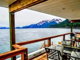 Covered porch with seating offers a view of a calm lake and snow-capped mountains.