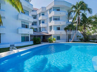 White apartment building with balconies overlooking a curved outdoor pool, surrounded by palm trees.