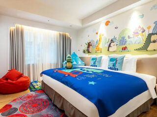 A children's bedroom with colorful animal-themed wall art, a blue bedspread featuring a dinosaur, and plush toys. There's a red bean bag and a patterned rug on the wooden floor.