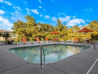 Outdoor swimming pool with several lounge chairs and two tables with orange umbrellas, surrounded by a metal fence. Trees are visible in the background under a blue sky.