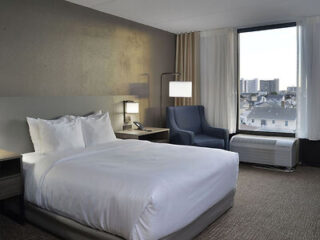 Hotel room with a neatly made bed, a blue armchair, and a large window with city views. Neutral-colored walls and carpet.