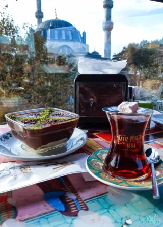 A cup of Turkish tea and a dessert with a sprinkle of green garnish are on a table, with a mosque visible in the background.